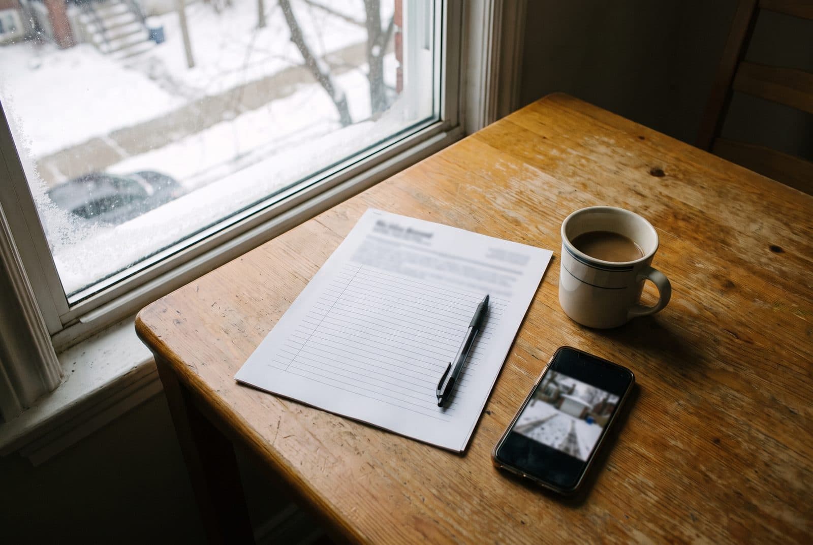 Quote sheet, pen, coffee and a phone showing a driveway photo on a table beside a snowy window Quote sheet, pen, coffee and a phone showing a driveway photo on a table beside a snowy window